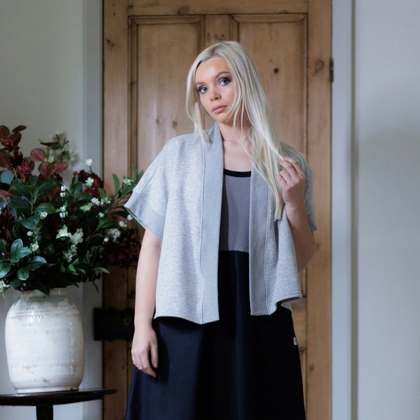 This is a young lady facing the camera wearing a grey marl shrug over a black and grey dress. She has long straight blonde hair which she twists with her left hand. Her right arm hangs loose by her side. There is a pot of flowers to the left and she is standing in front of an old pine wooden door.