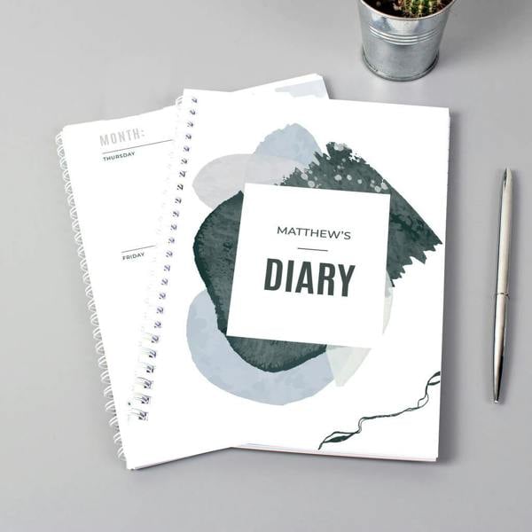 An open and closed personalised A5 softback diary featuring a blue abstract cover design printed with the name Matthew's Diary on a grey desk next to a pen.