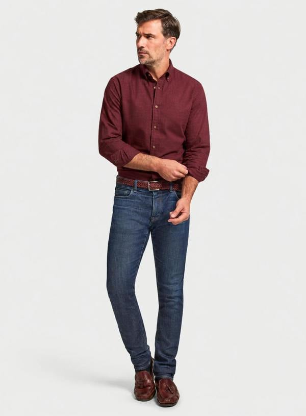 A man modeling a long-sleeve burgundy button-down shirt, dark blue jeans, a brown belt, and brown leather shoes against a light gray studio background.