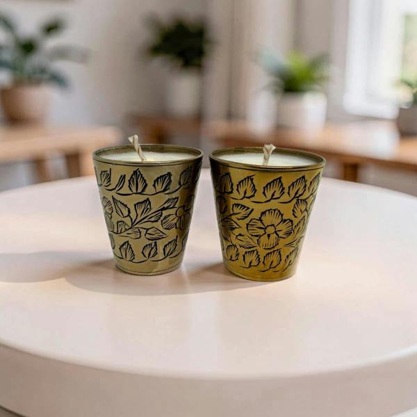 Two decorative metal candle holders with floral engravings, each containing a white candle, placed on a round white table in a softly blurred indoor setting.