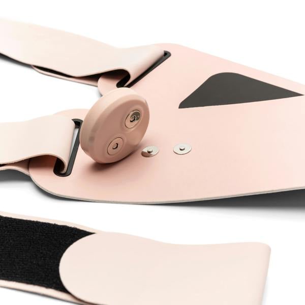 A close-up of the Ab, Tone & Shape light pink wearable electronic device with a round control button, snap fasteners, black accents, and Velcro details for muscle stimulation, displayed on a white surface.