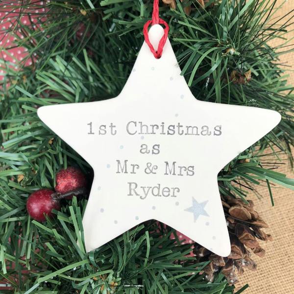 A white ceramic star ornament hanging on a Christmas tree branch, hand-stamped with the text '1st Christmas as Mr & Mrs Ryder' alongside a silver star motif.