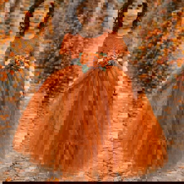 Young girl in an orange dress with a floral sash standing in a natural setting with autumn foliage.