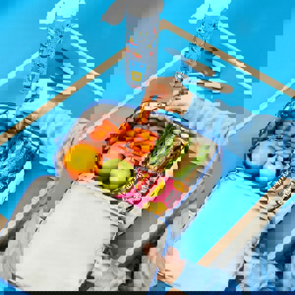 A packed lunchbox contains sandwiches, an apple, an orange, Walkers crisps, and carrot sticks. One hand picks up a carrot in a blue setting with a water bottle nearby.