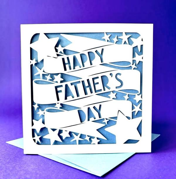 Stars Papercut Father's Day Card: A square white Father’s Day card featuring a laser-cut design of stars and flowing banners with the words "HAPPY FATHER'S DAY" stencilled out. The design reveals a blue background and sits atop a matching light blue envelope against a purple backdrop.