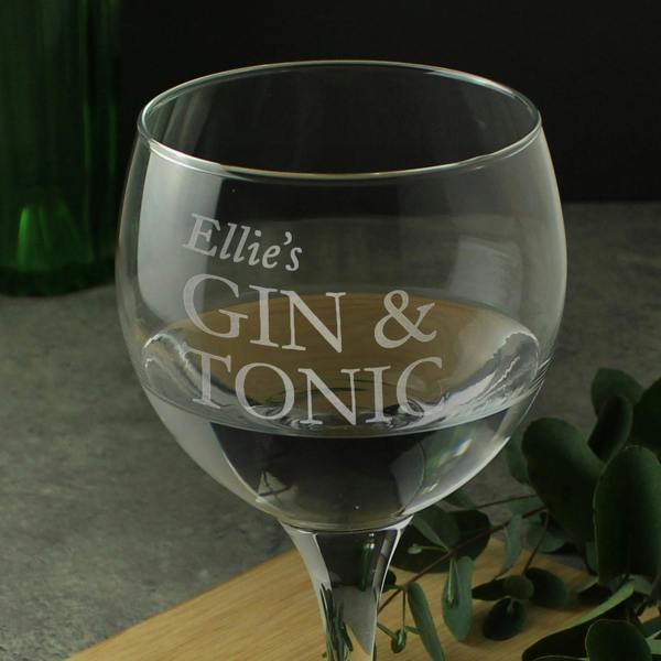 A close-up shot of a personalised stemmed gin balloon glass by Abode Home & Gifts, showing the frosted laser-engraved script name "Ellie's" above the fixed capital letters "GIN & TONIC", filled with a clear drink on a wooden serving board.