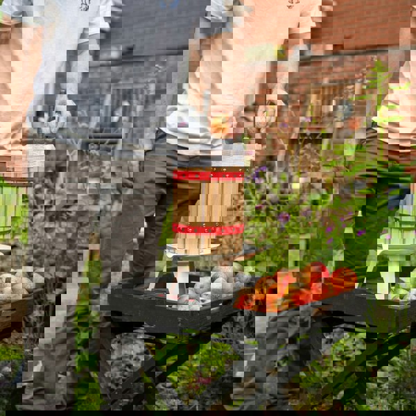 KuKoo 6 Litre Fruit Press with wooden barrel, red straps, and a person adding apples, set in a garden with fresh fruit nearby