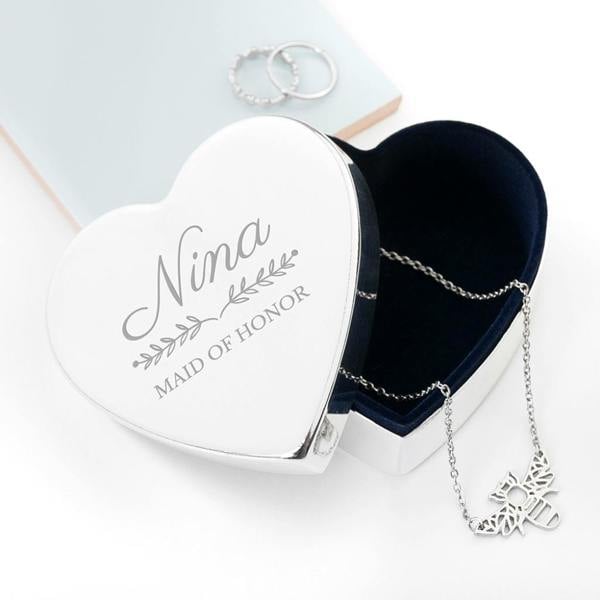 Top-down view of a heart-shaped silver jewellery box with a navy velvet lining. The lid is engraved with "Nina" in a script font and "Maid of Honor" in a serif font, separated by a delicate floral leaf design. A silver bee necklace is draped inside.