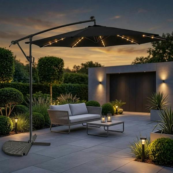 A large black 3m cantilever banana garden parasol with glowing solar LED lights along the interior ribs, shading a grey rattan sofa on a paved modern patio at sunset.