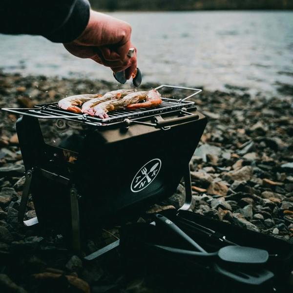 Hand using tongs to flip prawns on a Portable Notebook BBQ by a lake.