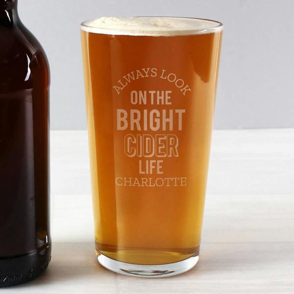 A personalised glass pint glass filled with golden cider and a white head, showcasing the engraved words Always Look On The Bright Cider Life and the name Charlotte next to a brown bottle.