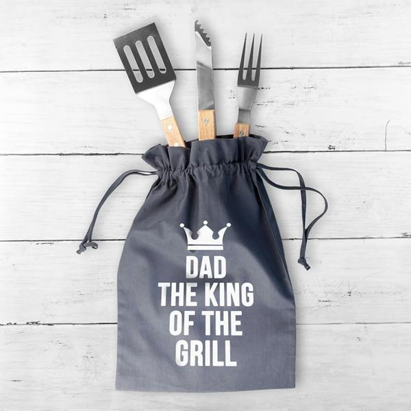 A slate-grey drawstring bag printed with a white crown and Dad The King Of The Grill text, with a stainless steel spatula, tongs, and fork tucked inside on a white wooden backdrop.