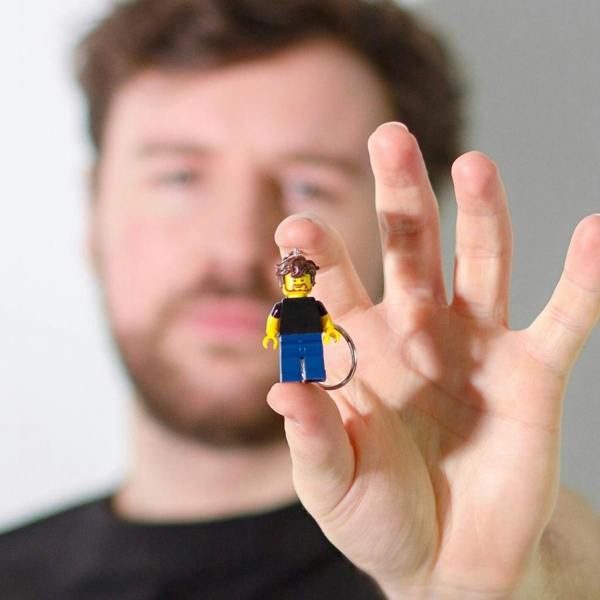 A close-up shot of a man's hand holding a tiny custom block figure keyring with brown hair, a bearded yellow face, a black t-shirt, and blue trousers, with the blurred face of the man matching the figure in the background.