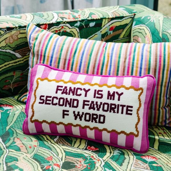 A small pink and white striped needlepoint cushion featuring the text "FANCY IS MY SECOND FAVORITE F WORD" propped on a colourful, patterned armchair.