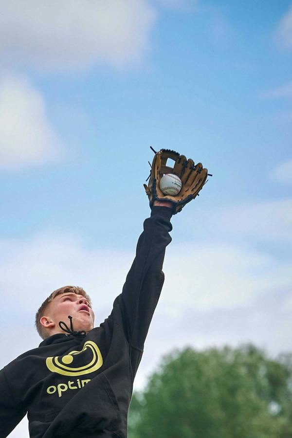Boy in black Optimum hoodie catching a white baseball in a brown mitt against blue sky - Optimum