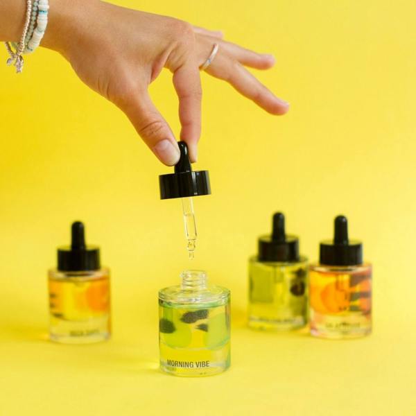 Hand holding a glass dropper above the Morning Vibe Daily Aromatherapy Mood Oil bottle surrounded by other mood oils against a bright yellow background by Abode Home & Gifts