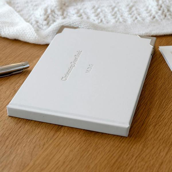 White Personalised Christening Guest Book with silver metal corners, foil-stamped with initials M.D.G on a wooden surface.