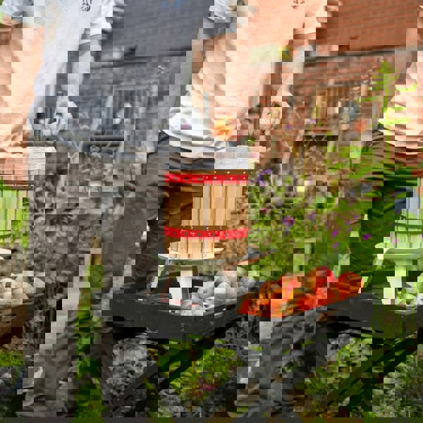 KuKoo 12 Litre Fruit Press with wooden barrel design, red straps, and a person adding apples, set in a garden with fresh apples nearby