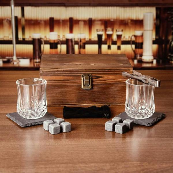 A dark wooden gift box with a metal clasp, surrounded by two crystal-style whisky tumblers on slate coasters, several granite chilling stones, and stainless steel tongs on a wooden bar top.