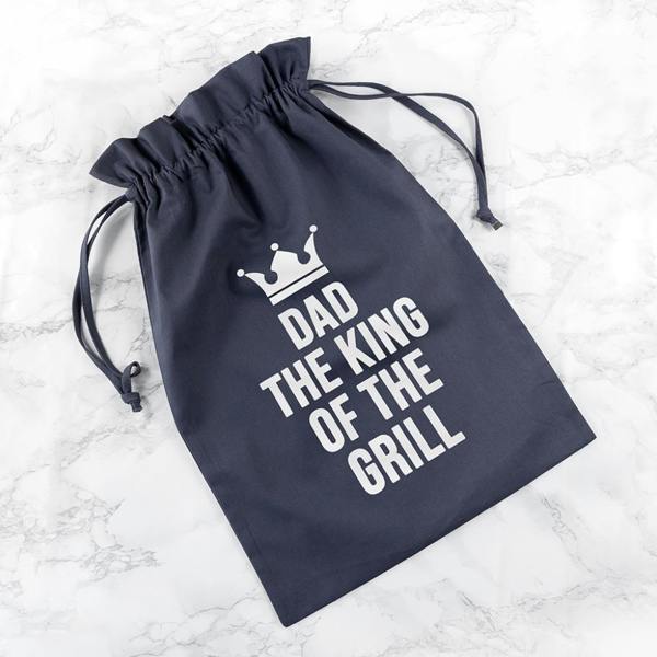 A slate-grey canvas drawstring pouch featuring a crisp white crown graphic and bold text reading Dad The King Of The Grill, displayed flat on a light marble background.