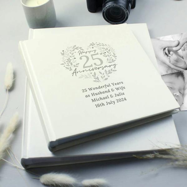 A cream-colored 25th Anniversary photo album resting on a gray surface, decorated with a silver heart-wreath design, personalized with '25 Wonderful Years as Husband & Wife', 'Michael & Julie', and '16th July 2024'.