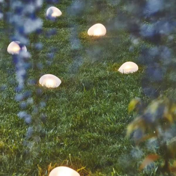 Multiple frosted glass solar pebble lights illuminating a garden lawn at twilight next to blooming purple wisteria trees.