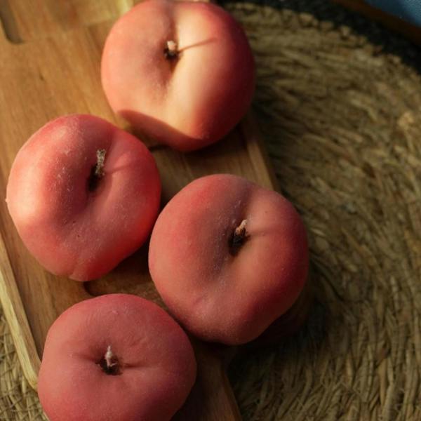 An overhead shot of four realistic, pink flat peach-shaped soy candles with a velvety finish laid out across a wooden paddle serving board.