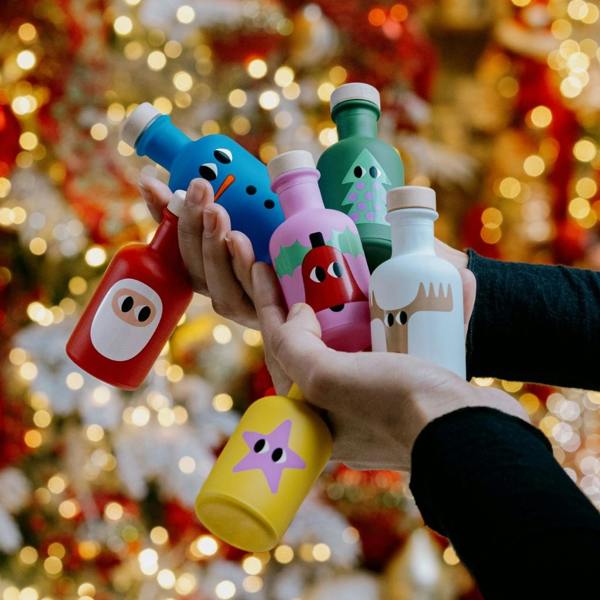 Hands holding six colourful miniature Christmas olive oil bottles decorated as festive characters, against a blurred background of warm golden fairy lights.