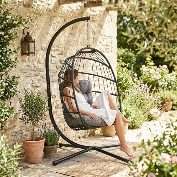 Black rattan hanging egg chair with plush gray cushions, suspended in a garden setting surrounded by flowers and greenery
