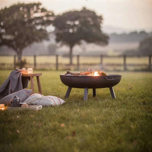 Statement cast iron garden fire bowl with a lit fire, shown in the large 100cm size against a scenic Cotswold-inspired countryside backdrop.