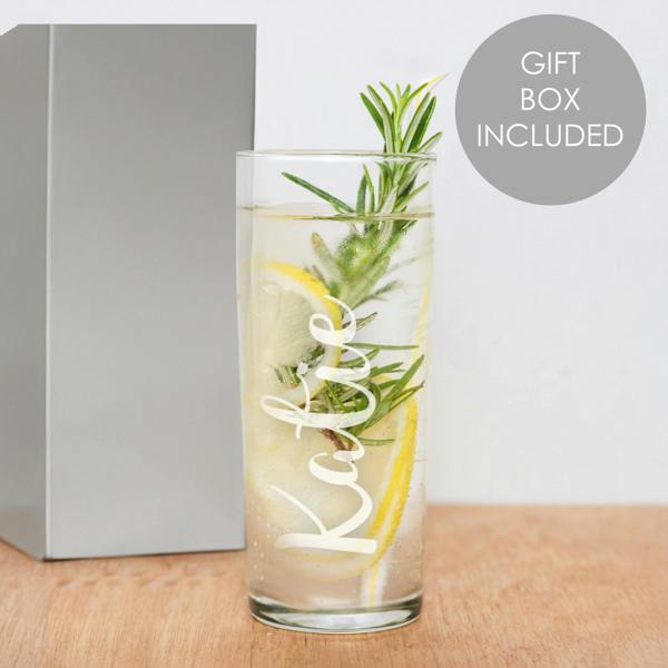 A clear glass hi ball tumbler filled with a clear carbonated drink, ice, lemon slices, and a fresh rosemary sprig sits on a wooden table. The name 'Katie' is engraved vertically on the front of the glass in a white script font. A grey folding gift box is visible in the background next to a circular 'Gift Box Included' graphic.