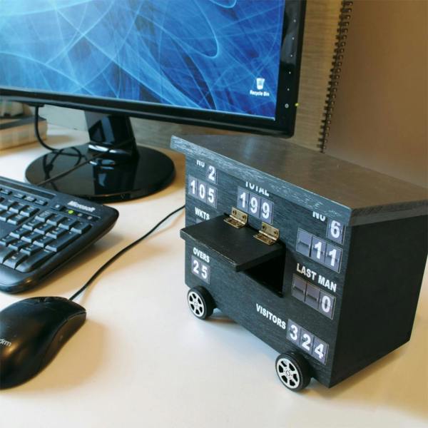 A wooden desk organiser designed to look like a black cricket scoreboard with movable panels, featuring "TOTAL", "WKTS", "OVERS", and "LAST MAN" text, sitting on a clean white desk next to a computer mouse and monitor.
