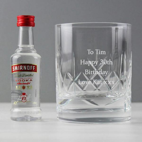 A heavy cut crystal spirit glass engraved with 'To Tim Happy 30th Birthday Love Kat xxx' standing next to a miniature 5cl Smirnoff vodka bottle.