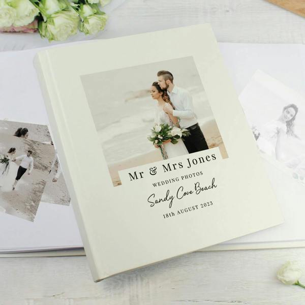 A cream hardback Personalised Photo Album reading Mr & Mrs Jones Sandy Cove Beach on the cover, featuring a wedding photo and resting on a white table next to white roses.