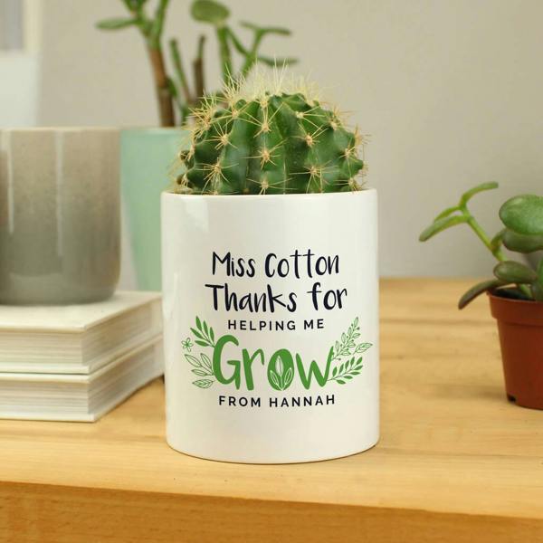 A white ceramic pot with a botanical design, featuring the text 'Mrs FISHER World's Best Teacher Love Polly xx', containing a green cactus on a wooden surface.
