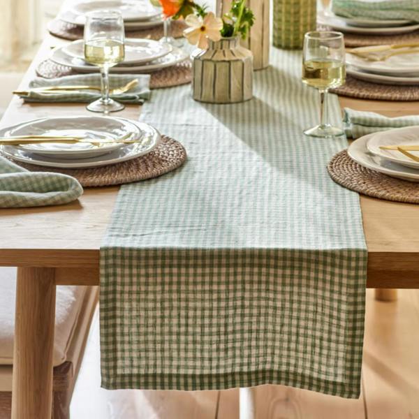 A rosemary green and cream checked linen table runner styled on an oak dining table with wine glasses and plates.