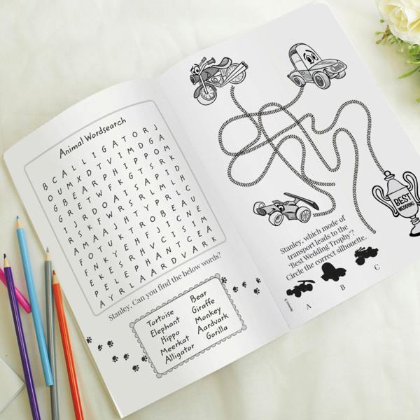 An open personalised children's wedding activity book shows two internal puzzle pages on a white background. The left page features an animal wordsearch matrix and reads 'Stanley, can you find the below words?'. The right page displays a vehicle tracking puzzle leading to a trophy and reads 'Stanley, which mode of transport leads to the Best Wedding Trophy?'. Colouring pencils lie to the left.