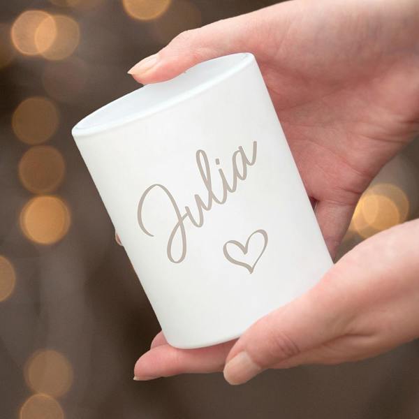 A close-up of hands holding a white frosted glass candle holder printed with the name 'Julia' and a small heart icon against blurred golden lights.