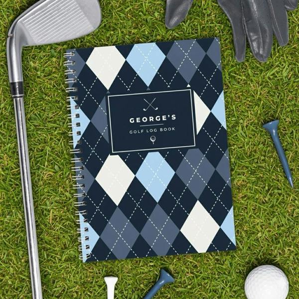 A personalised golf log book with a blue and navy argyle pattern bearing the name George's on the cover, laying flat on green grass alongside a golf club, ball, and tees.