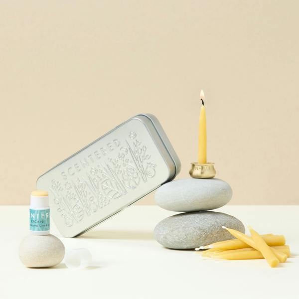 A relaxation balm set displayed with a metal tin, aromatherapy balm tube, a lit beeswax candle in a brass holder, and raw beeswax tapers.