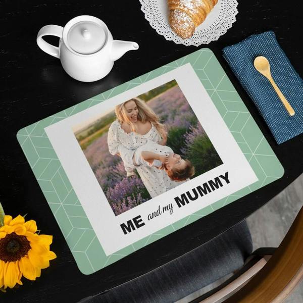 A high-angle view of the Mum's Personalised ‘Me And My Mummy’ Photo Placemat on a dark wooden dining table, set next to a white teapot, a croissant on a plate, a blue napkin with a wooden spoon, and a sunflower.
