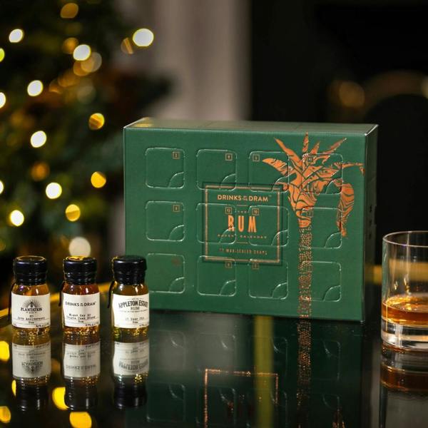 Green and copper foiled 12 dram rum advent calendar next to three wax-sealed miniature bottles and a glass of rum with Christmas tree lights.