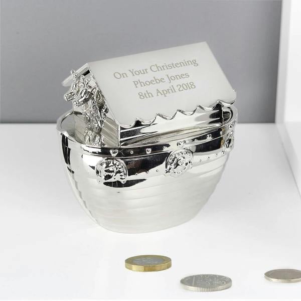 A high-shine silver nickel-plated Noah's Ark money box engraved on the roof with "On Your Christening Phoebe Jones 8th April 2018", sitting on a clean white surface with British coins scattered in front.