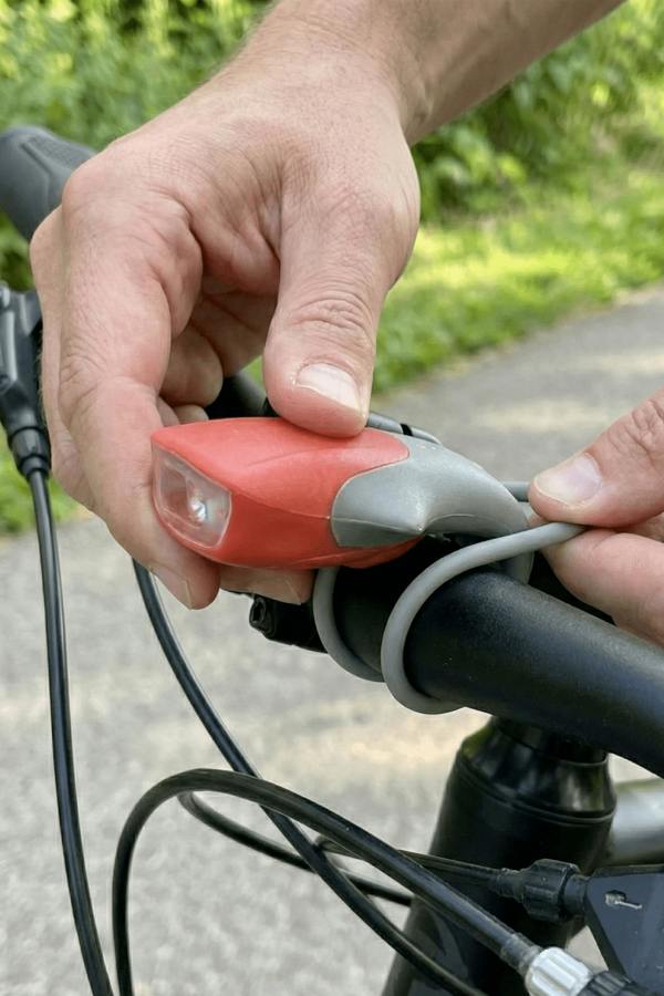 Hands fitting a red silicone LED bike light onto a handlebar with grey strap, outdoors - Optimum