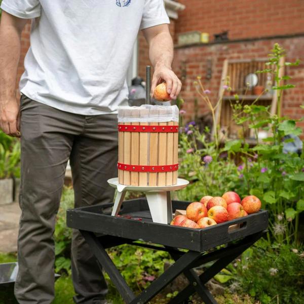 KuKoo fruit crusher with wooden barrel design, red accents, and a person adding apples, surrounded by fresh fruit in a garden setting