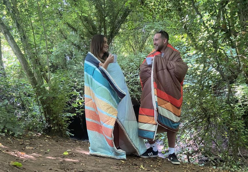 Tonal treetops and vivid spruce Insulated Outdoor Blanko been worn by a young couple enjoying the countryside with the outdoor blanket, wrapped around them. imageset:Vivid Spruce