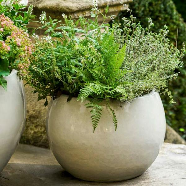 A medium-sized round white glazed ceramic planter filled with lush green ferns and herbs sitting on a stone patio step.