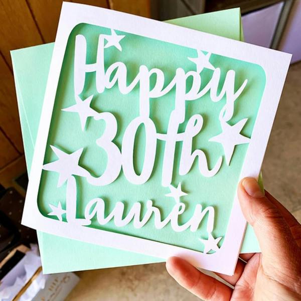 A hand holding a square, mint-green greeting card featuring a white, laser-cut cardstock overlay that reads "Happy 30th Lauren" surrounded by intricate star cut-outs.