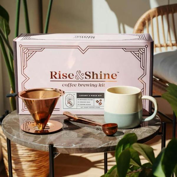 A Rise & Shine luxury coffee brewing kit featuring a rose gold pour-over filter, matching scoop, and beige and sea-green ceramic mug arranged on a round marble table in front of its gift box.