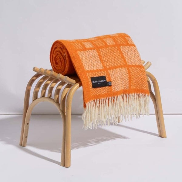 Bold orange wool blanket with white square patterns and fringed edges, neatly rolled on a wooden bench—luxury throw for indoor decor.
